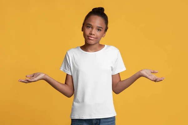 Confused teenager african american girl in white t-shirt makes choice, spread her arms to sides