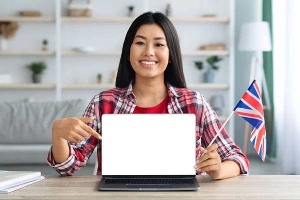 Aprenda inglês online. Mulher asiática com bandeira britânica apontando para laptop em branco — Fotografia de Stock