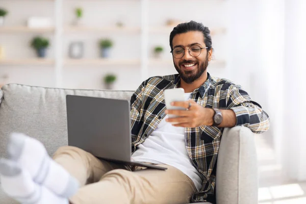 Indian guy sitting on sofa with laptop, holding smartphone