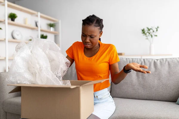 Bad delivery service, online shopping. Irritated black woman unboxing cardboard parcel, receiving damaged item at home