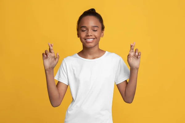 Happy calm teen african american girl in white t-shirt with closed eyes crossed fingers and makes a wish