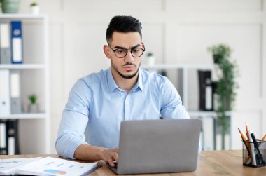 Endişeli genç bir Arap dizüstü bilgisayarla masasında oturuyor, hata yapıyor, son teslim tarihinden endişe ediyor.