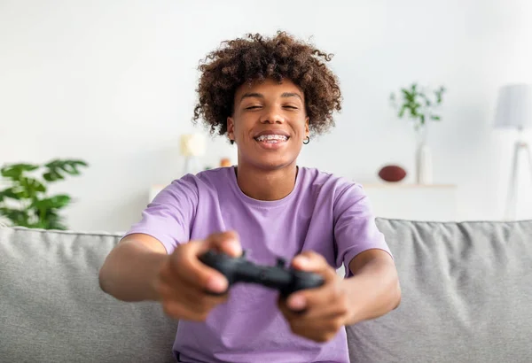 Entretenimiento doméstico. Joven adolescente negro alegre con joystick jugando juegos de ordenador en línea en el sofá en casa — Foto de Stock