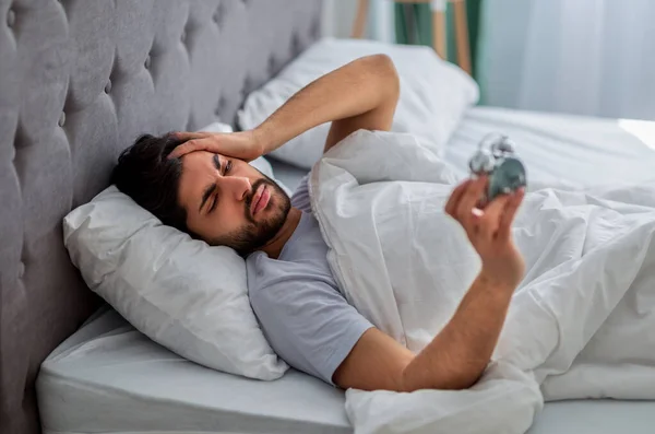 Hombre árabe infeliz acostado en la cama con despertador, no dispuesto a levantarse por la mañana y sufrir de dolor de cabeza — Foto de Stock