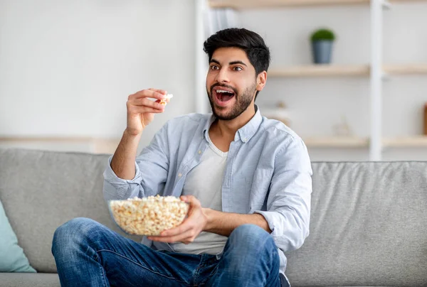 Retrato de homem árabe emocional assistindo TV e comer pipocas, descansando no sofá em casa, espaço livre — Fotografia de Stock