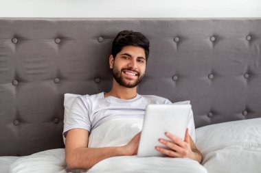 Hafta sonu eğlence konsepti. Mutlu Arap adam dijital tablet bilgisayar tutuyor, yatakta oturuyor, boşluğu kopyalıyor.
