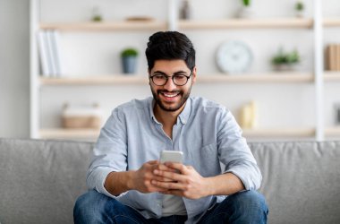 Gözlüklü mutlu Arap adam elinde akıllı telefonla oturuyor ve sosyal ağları tarıyor ya da mesajlaşıyor, boşluğu kopyalıyor