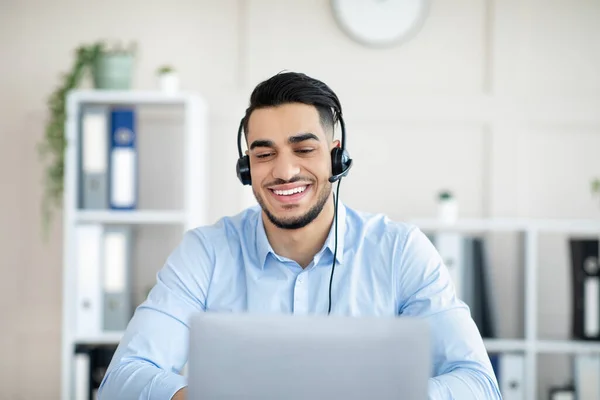 Hombre árabe alegre con auriculares que trabajan en línea en el ordenador portátil, teniendo videollamada en la oficina —  Fotos de Stock