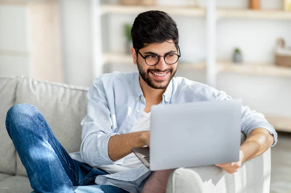 Feliz milenario árabe freelancer masculino trabajando desde casa, sentado en el sofá con el ordenador portátil, mirando a la pantalla y sonriendo — Foto de Stock