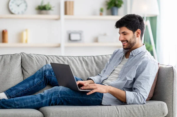 Gelukkig arabisch man rusten met laptop op de bank thuis interieur, kijken film of scrollen sociale netwerken — Stockfoto