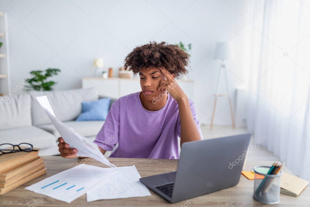 Bored irritated black teenager sitting at table with papers, using laptop to write coursework, studying online