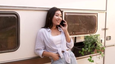 Genç Asyalı kadın modern karavanın yanında duruyor ve cep telefonuyla konuşuyor.