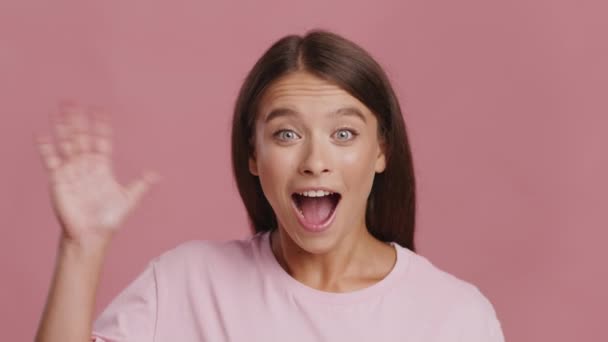 Возбужденная женщина машет рукой, улыбаясь в камеру на розовом фоне — стоковое видео