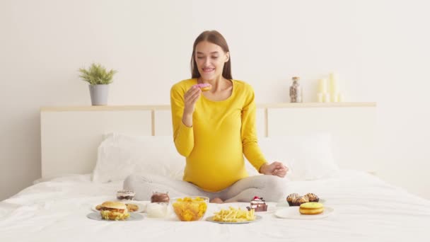 Caprichos alimentares de gravidez. Jovem grávida animada sentada na cama e desfrutando de doce donut lixo, câmera lenta — Vídeo de Stock
