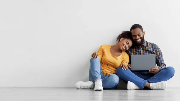 Веселый молодой черный мужчина и женщина смотрят в ноутбук, планируя интерьер, сидит на полу в пустой комнате — стоковое фото