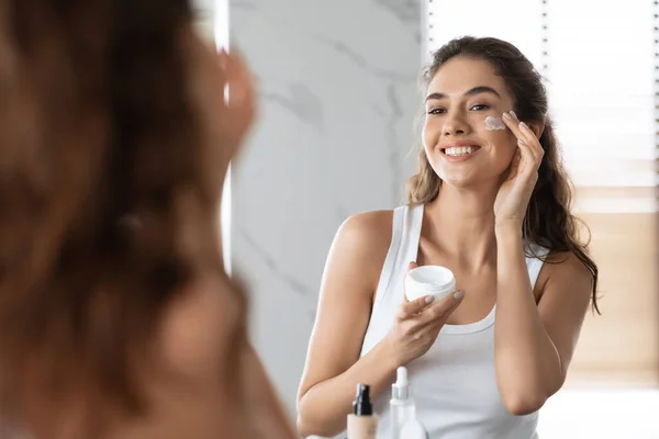 Giovane donna che applica crema sulle guance idratante pelle facciale all'interno — Foto Stock