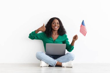 Dizüstü bilgisayarı ve Amerikan bayrağı olan siyahi kadın baş parmağını gösteriyor.