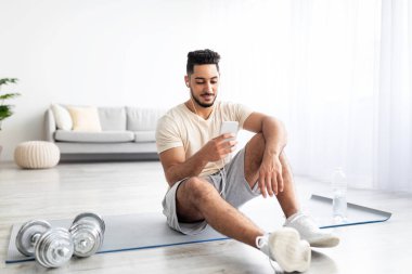 Ev içi spor eğitimi için müzik listesi seçen kulaklıkları ve cep telefonu olan yakışıklı genç bir Arap.