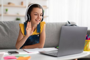 Bilgisayarı kullanan, webcam 'e el sallayan kulaklı bir kız.