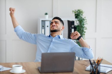 Bin yıllık Arap işadamı, iş yerinde ağrıyan kaslarını esnetiyor, modern ofiste dizüstü bilgisayarla oturuyor.