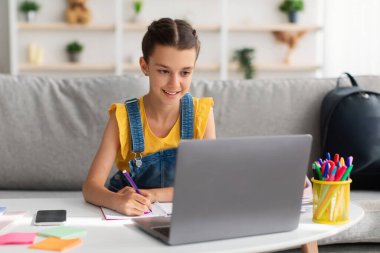 Neşeli kız masada oturuyor, dizüstü bilgisayar kullanıyor, defter çiziyor.