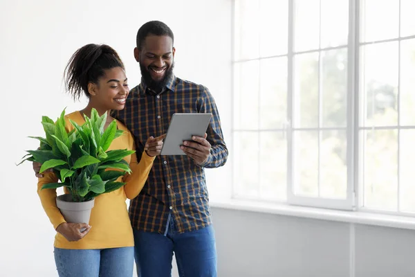 Heureux jeune couple afro-américain regardant en tablette, femme tenant plante, debout près de la fenêtre dans la chambre — Photo