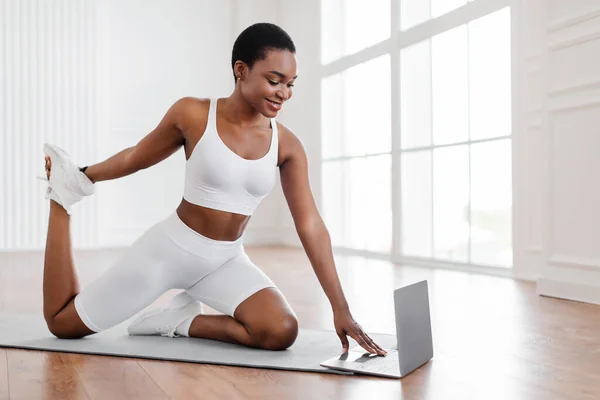 Веселая черная женщина растягивает мышцы ног с помощью персонального компьютера — стоковое фото