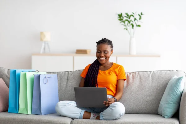 Afroamerikansk kvinna med bärbar dator köpa varor i nätbutik, sitter på soffan med shopper påsar, kopiera utrymme — Stockfoto