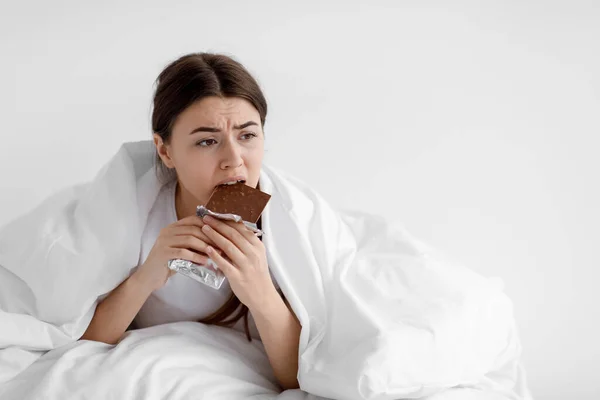 Smutna samotna głodna Europejka młoda dama jedząca czekoladę, owinięta w koc we wnętrzu sypialni — Zdjęcie stockowe
