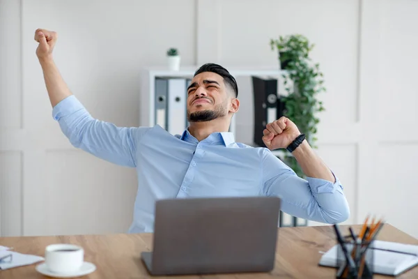 Hombre de negocios árabe milenario estirando sus músculos doloridos en el lugar de trabajo, sentado en el escritorio con computadora portátil en la oficina moderna — Foto de Stock