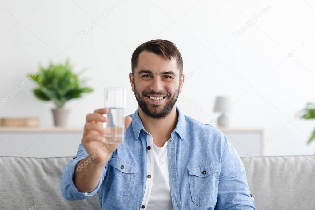 Smiling mature european male with beard shows glass of water in minimalist living room interior