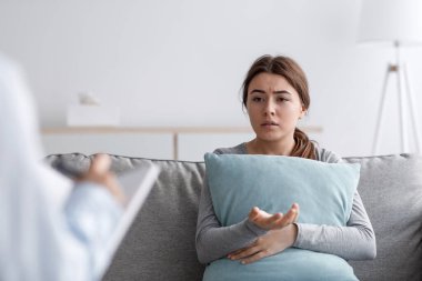 Üzgün mutsuz Avrupalı genç bayan kanepede oturuyor yastığa sarılıyor ve ofisteki psikologla konuşuyor.