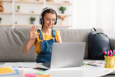 Neşeli liseli kız masada oturuyor, laptop kullanıyor, webcam 'e el sallıyor.