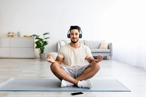 Jovem árabe cara com fones de ouvido e telefone celular ouvir música enquanto sentado em pose de ioga de lótus no tapete em casa — Fotografia de Stock