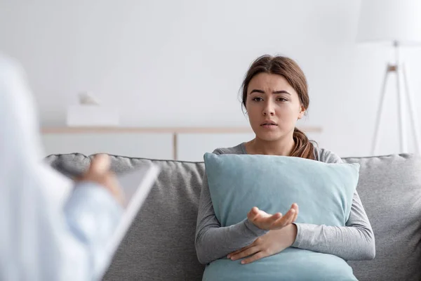 Triste triste disgustada joven europea sentada en el sofá abrazando almohada y hablando con el psicólogo en la oficina — Foto de Stock
