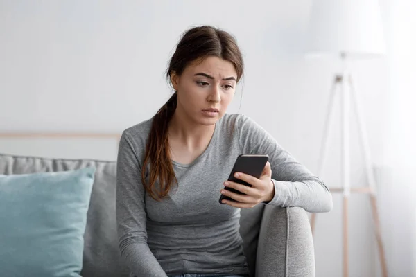 Сумна молода жінка сидить на дивані дивиться на смартфон і читає повідомлення вдома, розчарований отриманим сповіщенням — стокове фото