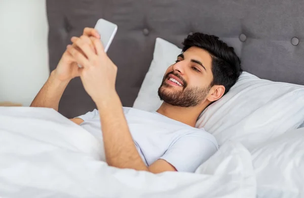 Redes sociales, buenas noticias matutinas. Feliz hombre árabe milenario despertando y mirando el teléfono inteligente, acostado en la cama — Foto de Stock