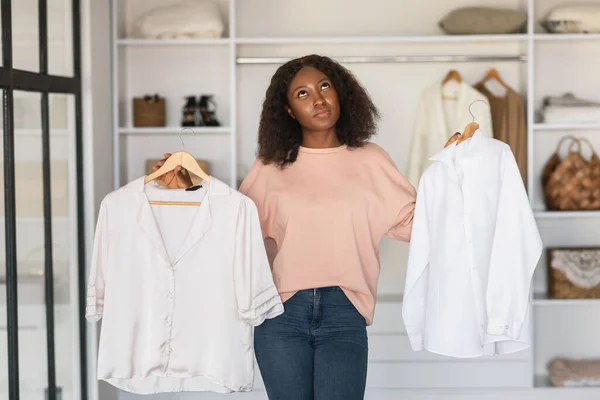 Pensive Black Woman Thinking Holding Two White Shirts At Home