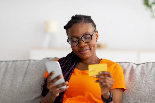 Härlig ung svart kvinna sitter på soffan, använder mobiltelefon och kreditkort, kollar ny app för online shopping — Stockfoto