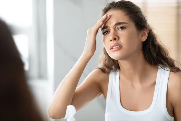 Ongelukkig jonge vrouw aanraken voorhoofd kijken naar rimpels in de badkamer — Stockfoto