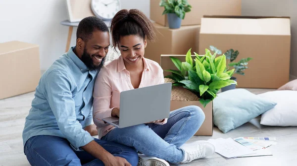 Sorrindo milenar Africano americano esposa mostrando ao marido laptop com novo design de quarto, sentado no chão com caixas — Fotografia de Stock