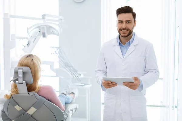 Hombre barbudo alegre dentista con tableta digital de pie por paciente —  Fotos de Stock