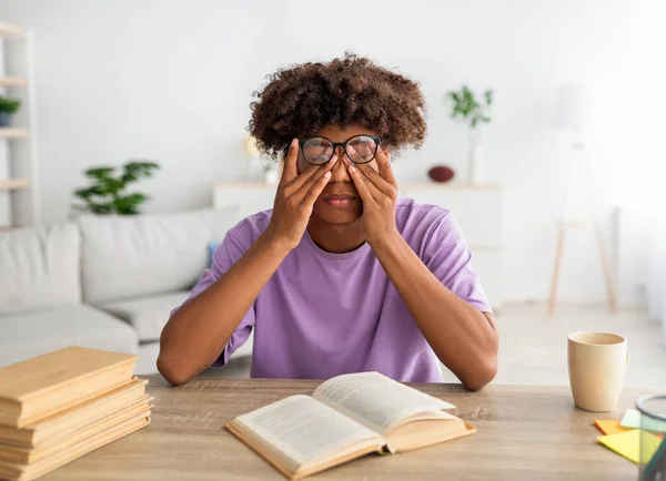 Czarny nastolatek facet tarcie zmęczony oczy siedząc przy biurko z otwarty podręcznik, wyczerpany z w domu edukacja — Zdjęcie stockowe