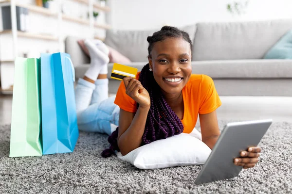 Bella signora nera sorridente sdraiata sul pavimento con tablet digitale, carta di credito e shopping bag a casa — Foto Stock