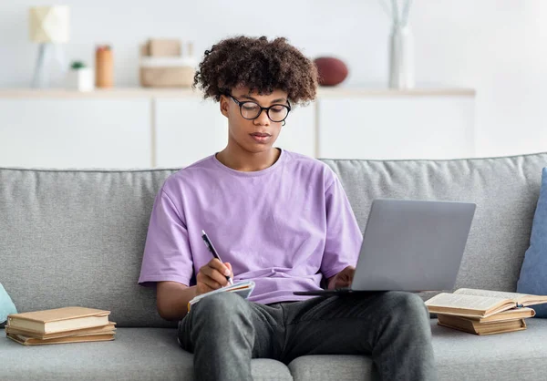 E-learning. Adolescente afro-americano focado tendo aula remota no laptop, tomando notas, sentado no sofá em casa — Fotografia de Stock