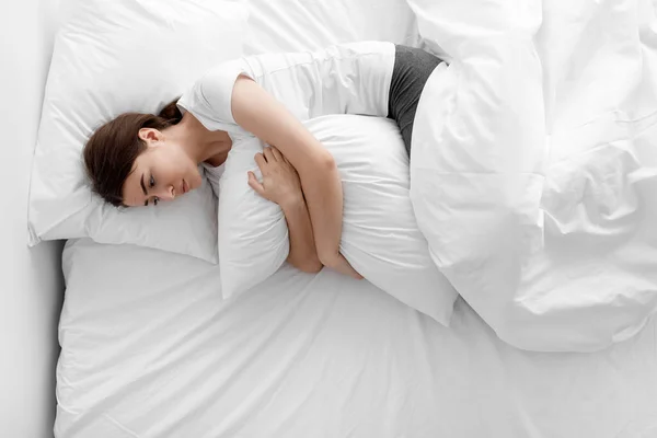 Zrozpaczona smutna Europejka millenijna przytulająca poduszkę i leżąca na białym łóżku w sypialni, widok z góry — Zdjęcie stockowe
