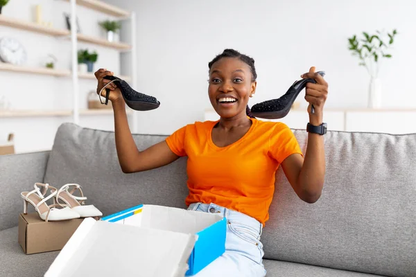 Zufriedenes Kundenkonzept. Positive schwarze Frau packt Karton aus, holt neue Schuhe aus Paket zu Hause — Stockfoto