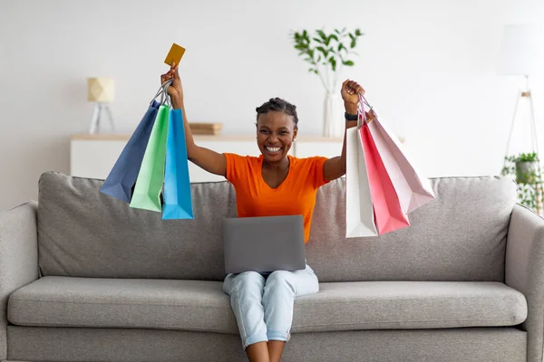 Excited Afro lady shopping online, sitting on couch with laptop and credit card, lifting gift bags above head at home