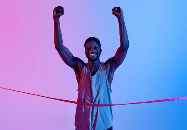 Esportista negro excitado correndo, cruzando a linha de chegada, levantando os braços em triunfo, celebrando a vitória na iluminação de néon — Fotografia de Stock