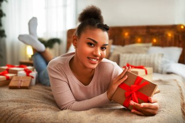 Pozitif Afro-Amerikalı kadın yatakta uzanıyor ve elinde Noel hediyesi kutusuyla kameraya gülümsüyor.